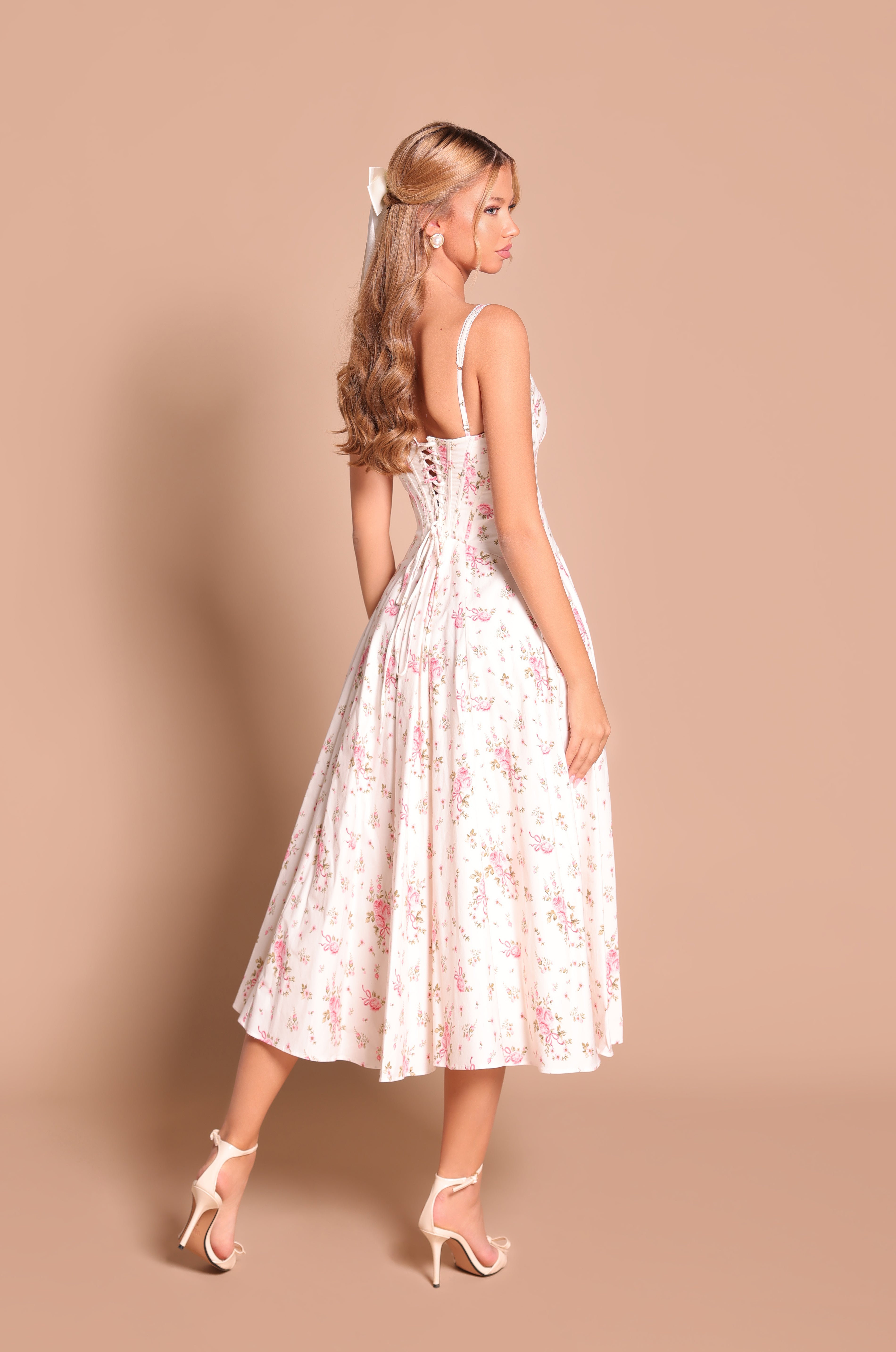 A woman in the Cream White Floral Casual Midi Dress: Rosella, featuring a fitted corset bodice and spaghetti straps, stands on a beige background, looking over her shoulder.